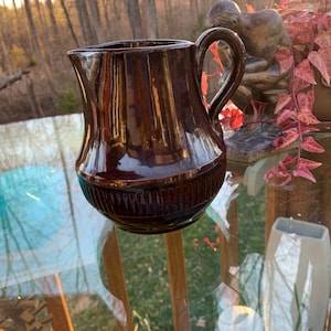 Puede incluir: Una jarra de cerámica marrón con asa. La jarra tiene un acabado brillante y un diseño acanalado alrededor de la base. La jarra está sobre una superficie de vidrio.