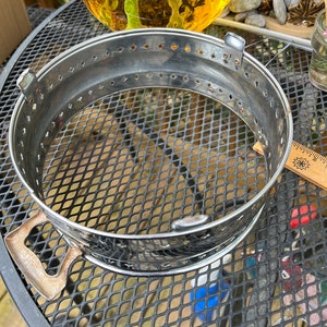 Puede incluir: Un anillo de metal plateado con un patrón decorativo de pequeños agujeros alrededor del borde. El anillo tiene dos asas en el lateral. El anillo está sobre una superficie de malla metálica negra.