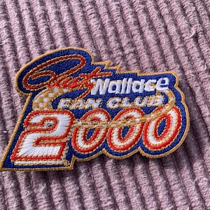 Puede incluir: Parche bordado con el texto "Rusty Wallace Fan Club 2000" en rojo, blanco y azul. El parche tiene un borde dorado y está sobre una superficie rosa texturizada. El diseño está en una fuente estilizada.