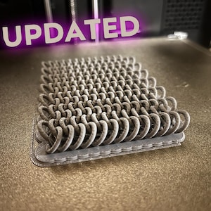 May include: A 3D printed gray rectangular object featuring a repeating interlocking coil pattern on a flat base. The word UPDATED is displayed in white text with a purple glow above the object, which sits on a textured metallic print bed.