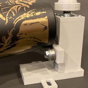 May include: A white 3D printed rotating stand with a black and gold cylinder attached. The stand has a base with a small rectangular extension. The cylinder has a gold design on a black background.