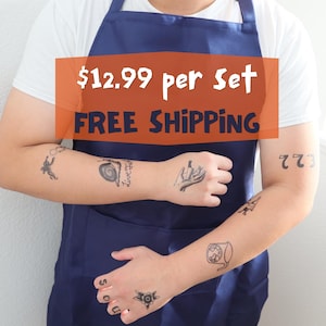 May include: A person wearing a white t-shirt and a navy blue apron, with temporary tattoos on their arms and hands. The tattoos include various small black illustrations and the number "773".