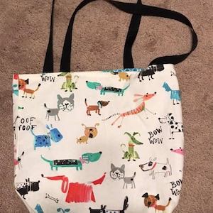 May include: A white canvas tote bag with black straps. The bag is decorated with a colorful pattern of dogs and the words "Woof", "Bow Wow", and "Wow".