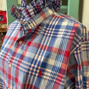 May include: A long-sleeved button-down shirt featuring a red, white, and blue plaid pattern. The shirt has a high, ruffled collar and a small red embroidered logo on the chest. The front is secured with white buttons down the center placket.