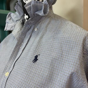 May include: A blue and white checkered shirt with a ruffled collar. The shirt features a small navy blue logo on the chest. The shirt is buttoned at the collar and has a classic, preppy style.