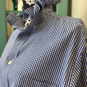 May include: A long-sleeved button-down shirt featuring a blue and white gingham check pattern. The shirt includes a high, ruffled collar detail and a single chest pocket on the left side. The garment is displayed on a black mannequin torso.