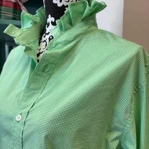 May include: A light green and white gingham button-down shirt featuring a high ruffled collar. The shirt is constructed with a standard placket, white buttons, and long sleeves, showcasing a classic checkered pattern throughout the fabric.