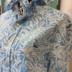May include: A light blue button-down shirt featuring a white tropical leaf pattern, a ruffled collar, and a chest pocket with an embroidered whale logo. The shirt has a front button placket with light-colored buttons and long sleeves.