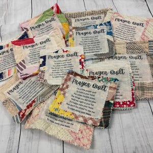 May include: A collection of handmade prayer quilt squares with different fabrics and designs. Each square features a prayer printed on it. The squares are arranged in a pile on a white wooden surface.