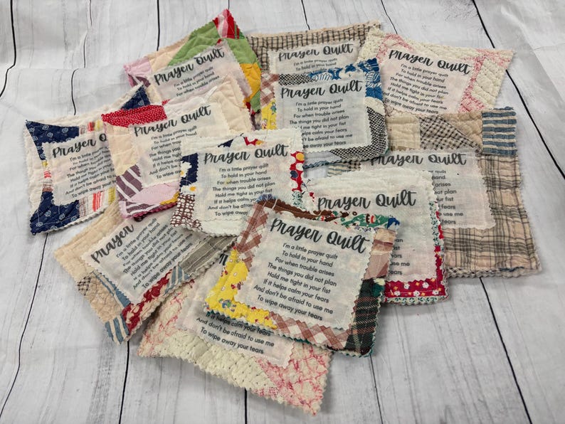 May include: A collection of handmade prayer quilt squares with different fabrics and designs. Each square features a prayer printed on it. The squares are arranged in a pile on a white wooden surface.