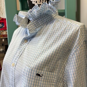 May include: A light blue and white gingham button-down shirt featuring a ruffled collar and a chest pocket with a small embroidered whale logo. The shirt has a front button placket with light-colored buttons and long sleeves, displayed on a mannequin.