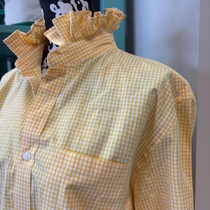 May include: A long-sleeved button-down shirt featuring a yellow and white gingham check pattern. The shirt includes a high, ruffled collar, a front button placket with white buttons, and a single chest pocket on the left side of the garment.