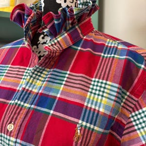 May include: A red plaid shirt with a ruffled collar. The shirt features a button-down front and a small embroidered logo on the chest. The plaid pattern includes red, blue, green, yellow, and purple lines.