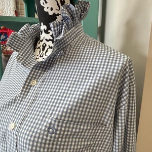 May include: A long-sleeved button-down shirt featuring a blue and white gingham check pattern. The shirt includes a ruffled collar, a front button placket with white buttons, and a single chest pocket with a small circular embroidered logo.