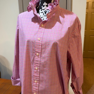 May include: A long-sleeved button-down shirt featuring a pink, green, and white micro-check pattern. The shirt has a high, ruffled collar and is displayed on a mannequin with a black and white patterned neck piece. The sleeves are rolled up.