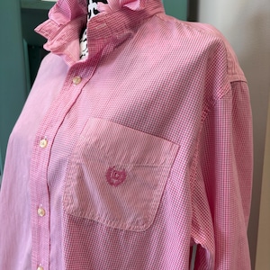 May include: A long-sleeved button-down shirt in a pink and white gingham pattern. The shirt features a ruffled collar, a front button placket with light-colored buttons, and a single chest pocket with an embroidered logo in pink thread.