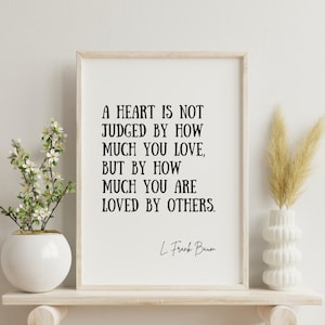 May include: A framed print with the quote "A HEART IS NOT JUDGED BY HOW MUCH YOU LOVE, BUT BY HOW MUCH YOU ARE LOVED BY OTHERS" by L. Frank Baum. The print is displayed on a wooden shelf with a white vase and decorative plants.