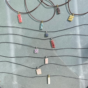 May include: A collection of necklaces with various pendant designs. The pendants are rectangular, square, and irregular shapes in colors like red, blue, gold, and purple. The necklaces have black cords and brown leather cords.