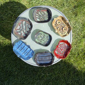 May include: A collection of seven ceramic soap dishes in various shapes and colors, including blue, red, green, black, and tan. Each dish features unique cut-out patterns or drainage holes designed to hold soap bars, arranged on a circular surface.