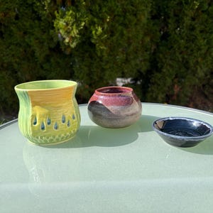 May include: A set of three ceramic pieces including a lime green and yellow cup with a textured raindrop pattern, a small rounded bowl with a red and grey gradient glaze, and a shallow black dish with a glossy finish.