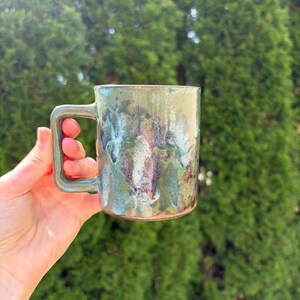 May include: A handmade ceramic mug with a square handle. The mug has a mottled glaze in shades of green, blue, and brown. The mug is held in a hand against a backdrop of green foliage.