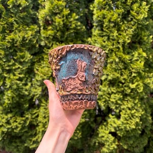 May include: A decorative ceramic planter with a textured, earthy design. The planter features a relief of a rabbit and foliage, with a turquoise interior. The exterior has a brown and green color scheme, perfect for small plants or succulents.