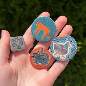 May include: A collection of handmade ceramic pins featuring animal designs. Includes a gray square pin with a bat, a blue pin with an orange cat, a red pin with a black cat, and a blue pin with a bird. The cat pin has the word "mao" on it.