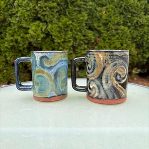 May include: Two ceramic mugs with square handles and unglazed terracotta bases. The left mug features a blue and green swirled glaze, while the right mug displays a black, blue, and tan swirled glaze pattern. Both mugs have a cylindrical shape.
