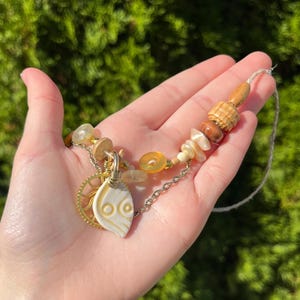 May include: Handmade necklace featuring a cream-colored, leaf-shaped pendant with two eye-like designs. The necklace includes a gold-colored gear charm, wooden beads, and a variety of other beads in shades of cream, brown, and yellow. The necklace is held in a hand.