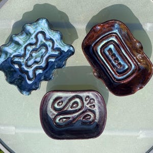 May include: Three handcrafted ceramic dishes in shades of blue, brown, and purple. The dishes have unique, organic shapes and textured surfaces. The blue dish has a wavy edge, the brown dish is rectangular, and the purple dish has a swirling design.