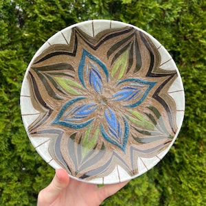 May include: A ceramic plate with a white background and a brown, green, and blue floral design. The design features a large flower with multiple petals.