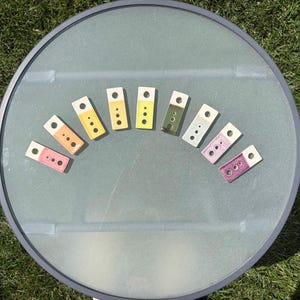 May include: Nine rectangular ceramic test tiles arranged in a semi-circle. Each tile features a white top section with a single hole and a glazed bottom section in varying colors including pink, orange, yellow, lime, green, blue, and purple.