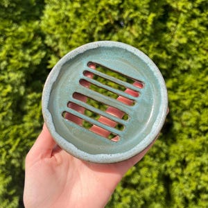 May include: A round, ceramic soap dish with a turquoise glaze and a series of parallel slots. The dish has a slightly irregular edge and a light blue rim. The background is a blurred green.