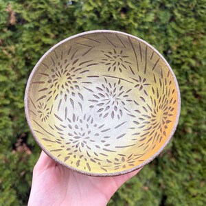 May include: A handmade ceramic bowl with a yellow and brown color scheme. The bowl features a floral pattern etched into the surface. The interior has a gradient of yellow hues, and the rim is a darker brown.