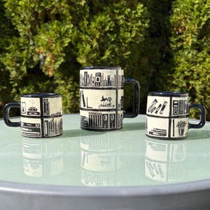 May include: Three ceramic mugs with a cream-colored base and black hand-painted illustrations of bookshelves, books, and potted plants. Each mug features a square handle and a grid-like pattern depicting various library-themed items.