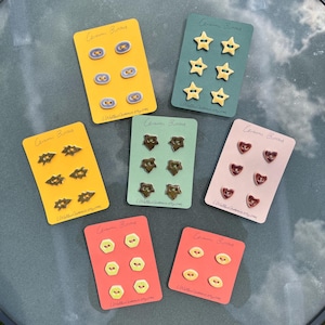 May include: A collection of ceramic buttons in various shapes and colors, including stars, hearts, and ovals, displayed on colorful cards. The cards are labeled "Ceramic Buttons" and feature a website address.