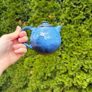 May include: A small, round ceramic teapot with a vibrant blue speckled glaze. The teapot features a curved handle, a short spout, and a matching lid with a small knob. The base and rim of the teapot show a dark brown, unglazed clay texture.