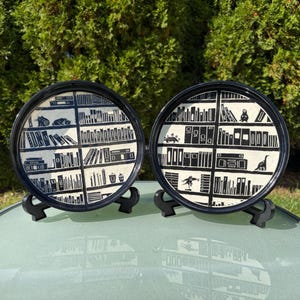 May include: Two round ceramic plates with black rims, each featuring a hand-painted black and white illustration of a multi-tiered bookshelf filled with various books, small cat figures, and dinosaur silhouettes on a cream background.
