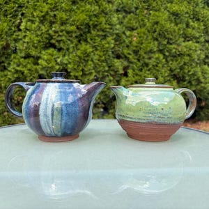 May include: Two ceramic teapots with lids. The left teapot features a vertical drip glaze pattern in shades of blue, purple, and white. The right teapot has a light green glaze on the upper half and a raw, unglazed reddish-brown clay base.