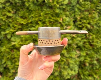 May include: A ceramic tea strainer with a cylindrical body featuring a band of small circular perforations. The piece has a smooth, dark grey glazed finish on the top and bottom sections, with a natural tan clay texture in the middle.