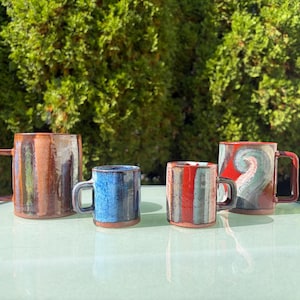 May include: Four ceramic mugs of varying sizes and shapes. The first is tall with brown and blue vertical stripes. The second is a small blue mug. The third is a small mug with red and grey stripes. The fourth is a wide mug with red and blue swirls.