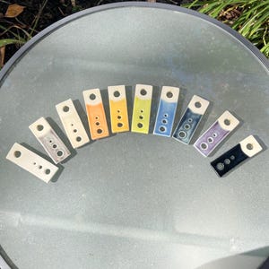 May include: A collection of rectangular ceramic pendants in various colors, including white, orange, yellow, green, blue, purple, and black. Each pendant has a hole at one end and a series of small, circular indentations. The pendants are arranged in a semi-circle.