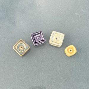 May include: Four square ceramic beads in shades of yellow, purple, and blue-green. Each bead has a central hole and a raised square design. The beads are arranged on a gray surface.