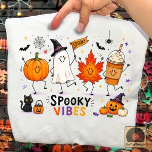May include: A white t-shirt featuring a colorful Halloween graphic with a smiling pumpkin, a ghost in a witch hat holding a Spooky flag, an orange maple leaf, and a coffee cup. The text Spooky Vibes is printed below the characters.