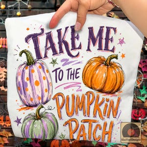 Puede incluir: Una camiseta blanca con un estampado gráfico que incluye el texto "TAKE ME TO THE PUMPKIN PATCH" en morado y naranja. El diseño muestra tres calabazas: una morada con puntos naranjas, una naranja y una verde con rayas moradas.