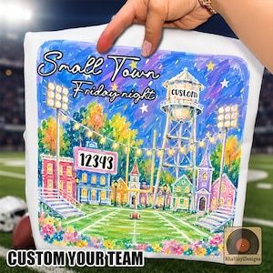 May include: A white t-shirt featuring a colorful painterly illustration of a football field at night. The design includes a water tower labeled custom, a scoreboard with the number 12343, and the text Small Town Friday night.