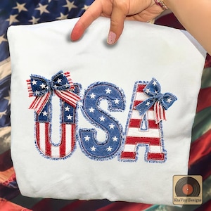 May include: A white t-shirt featuring the letters USA in a patriotic design. The U and A are decorated with red and white stripes and blue star patterns, while the S is blue with white stars. Two denim-style bows are attached to the letters.