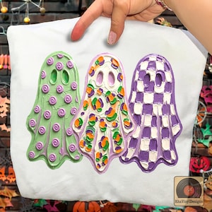 May include: A white t-shirt featuring three ghost illustrations with textured paint effects. The left ghost is light green with purple swirls, the center ghost has a white base with orange and green spots, and the right ghost has a purple and white checkerboard pattern.