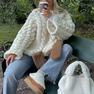 Op de afbeelding: Crèmekleurige, grofgebreide trui met een kabelpatroon. De outfit omvat lichtblauwe jeans, witte plateau laarzen met een bruine zool en een fluffy witte tote bag. Een kleine bruine tas en bijpassende wanten maken de look compleet.