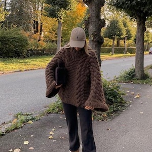 May include: A person wearing a brown cable-knit sweater with wide sleeves, black pants, a tan baseball cap, and brown shearling-lined slippers. The outfit is worn outdoors on a paved path with trees in the background.